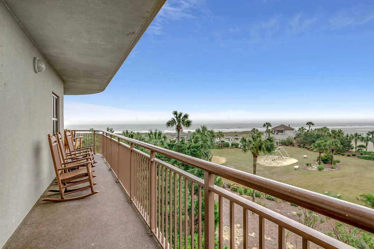 An inviting exterior scene featuring rocking chairs arranged on a patio, overlooking a beautifully landscaped garden with palm trees and a stunning ocean view beyond. The area is bathed in natural light, creating a serene, welcoming atmosphere that encourages relaxation and socializing. Children can be seen playing in the garden, adding an element of family fun.