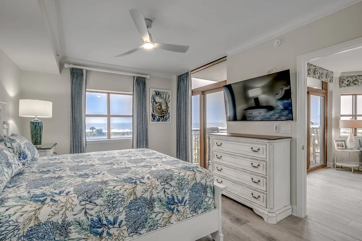 A tranquil bedroom with a bed adorned in floral bedding, a large window offering expansive ocean views, tasteful decorative artwork, and a sleek wall-mounted television. Warm lighting and a restful atmosphere are evident.