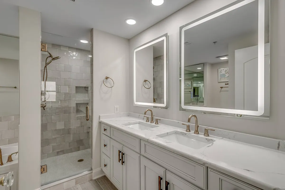 A modern, well-lit bathroom featuring a double sink vanity, large mirrors, and a glass shower enclosure. The setting is luxurious and designed for convenience, supporting rejuvenating routines and comfortable group stays.