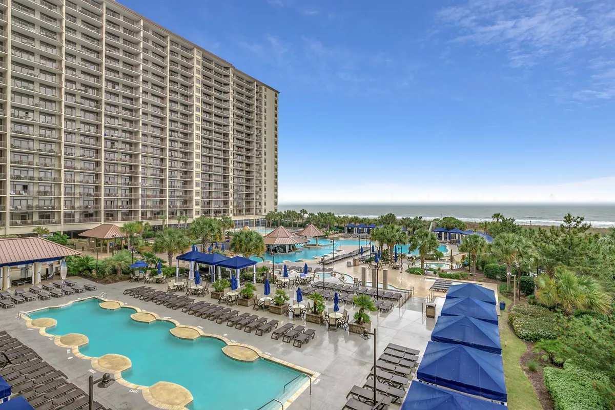 The image showcases an expansive pool area framed by swaying palms and lush tropical landscaping, with inviting blue loungers and umbrellas set against the backdrop of the sparkling Atlantic Ocean. The scene captures the golden hues of sunset, suggesting a perfect space for family relaxation, poolside gatherings, and enjoying the coastal breeze just steps from the sand.