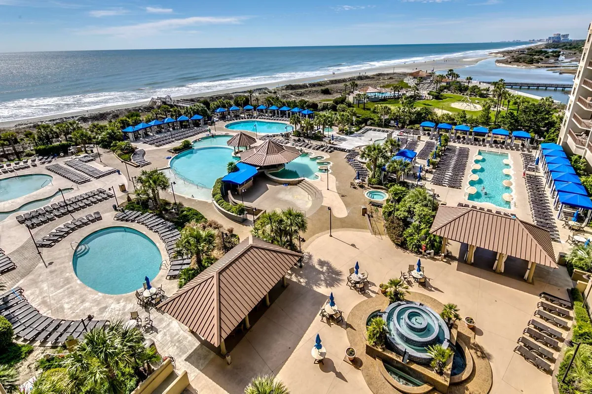 A sweeping view of the resort's outdoor swimming pools with glistening blue water, surrounded by palm trees, cabana structures, and lounging areas. The backdrop includes the ocean, enhancing the sense of a true beach resort getaway. The scene is lively yet serene, perfect for relaxation and socializing.