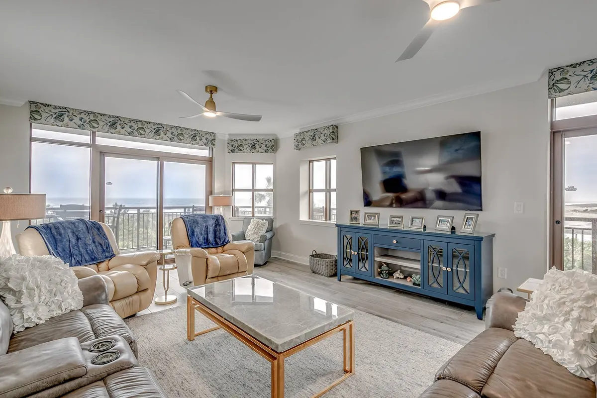 A cozy, tranquil living room furnished with plush blue seating, a large TV, and expansive windows that frame stunning ocean views. The natural light floods the space, highlighting the inviting decor and the seamless blend between comfort and coastal sophistication.