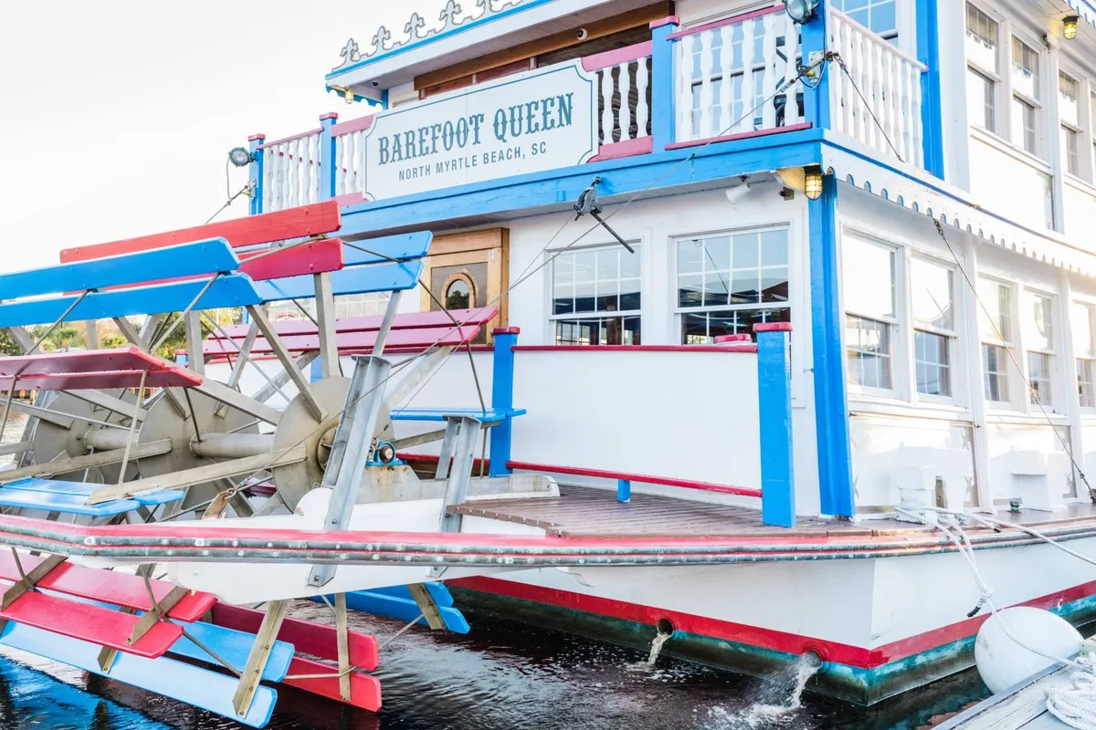 A colorful paddlewheel riverboat docked by the water, adorned with signage and details hinting at dining and live entertainment. The inviting exterior suggests a unique dinner cruise experience with sunset views.