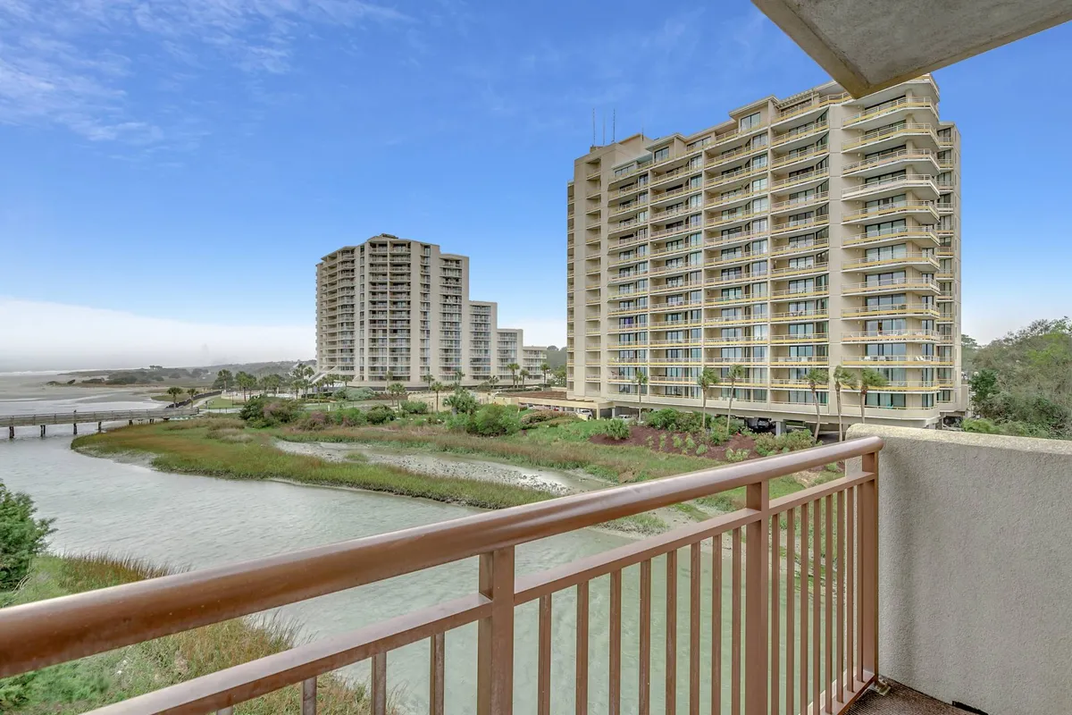 A balcony with a view over water and surrounding lush greenery, with buildings visible in the distance. The balcony railing establishes a sense of privacy while the expansive view connects guests to nature and the outdoors, offering a glimpse of the broader resort and its beach access.