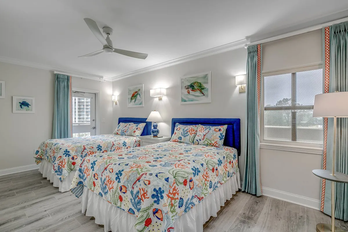 A bright, inviting guest bedroom featuring two beds with colorful bedspreads, decorative wall art, a nightstand with a lamp, and natural light streaming through the window. The mood is fresh and welcoming, ideal for families or groups.