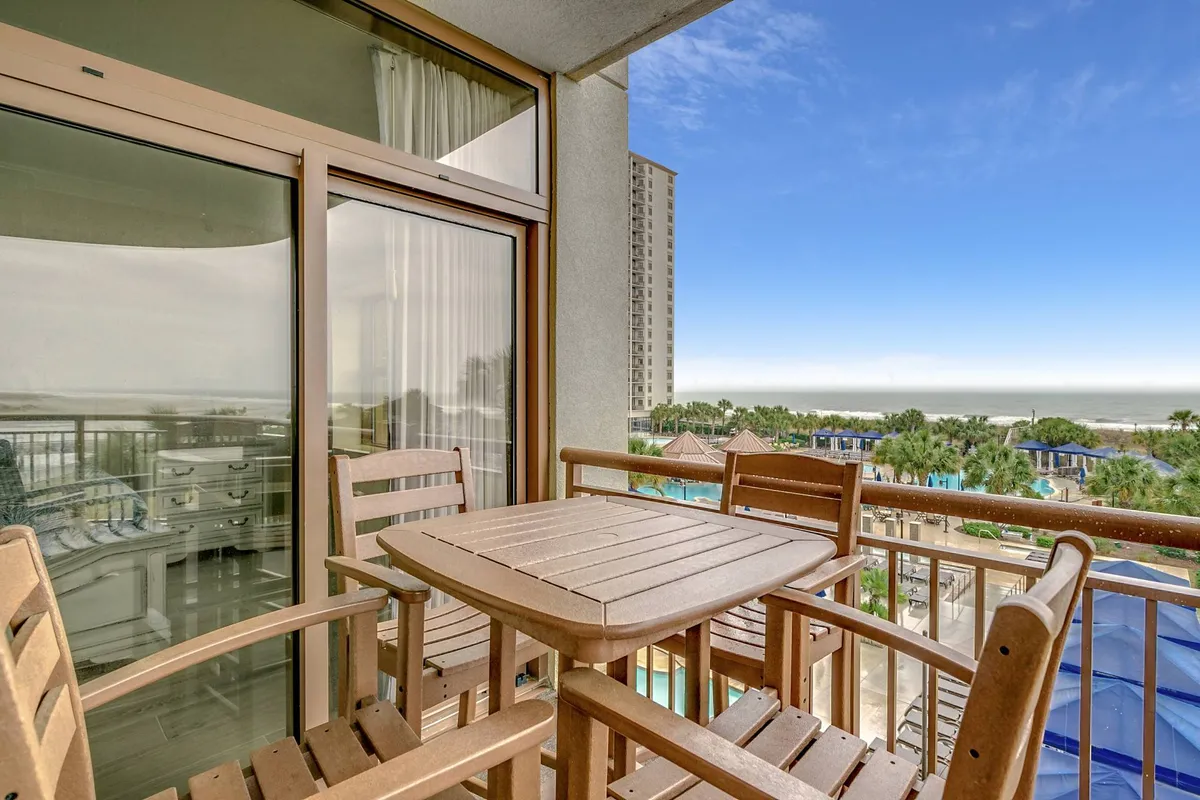 A peaceful balcony scene featuring wooden furniture overlooking the sparkling ocean and the resort’s pool area below. The view extends across the coast, with ample seating inviting guests to unwind while taking in both sunrise and sunset vistas.