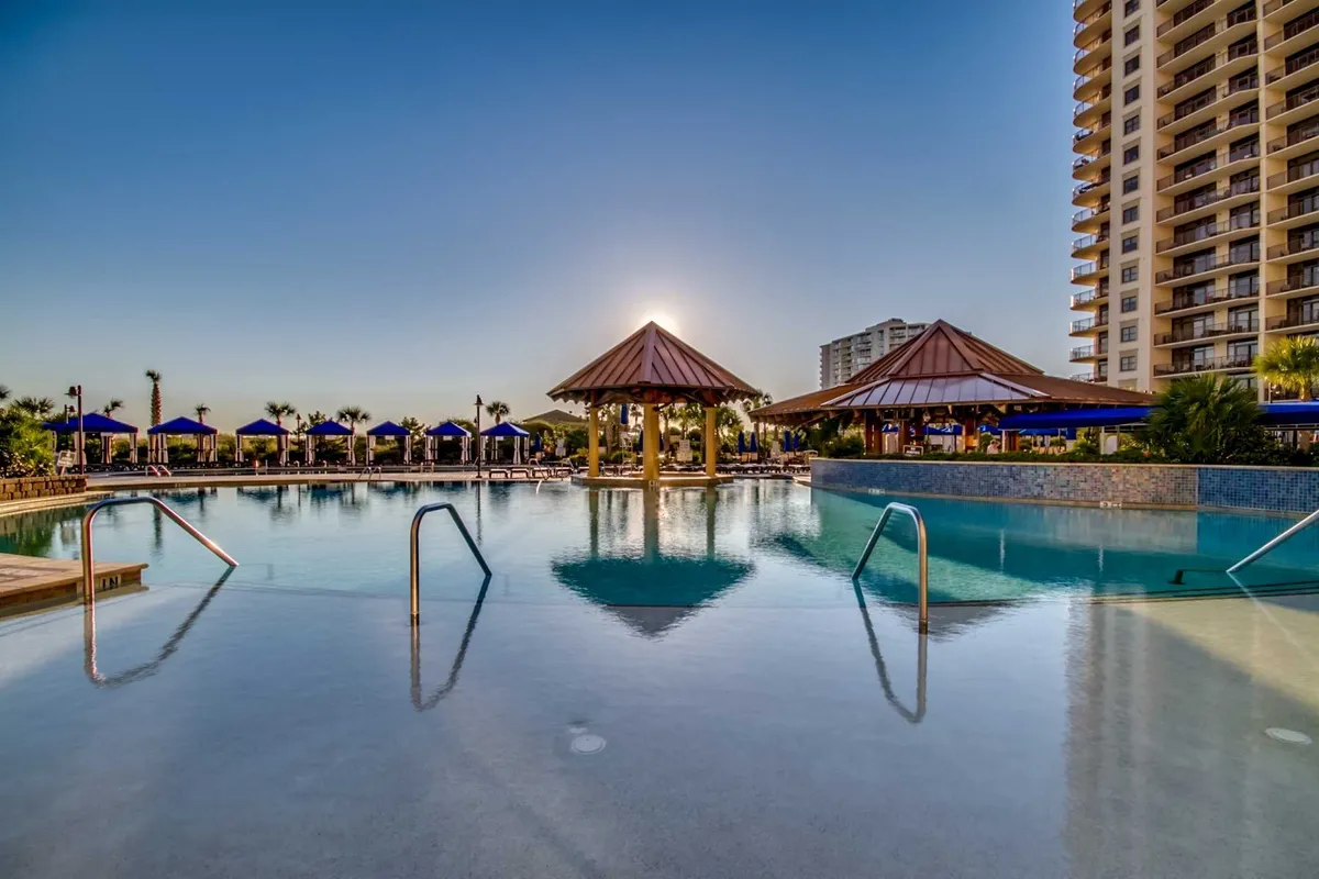 A luxurious resort pool complex with sparkling blue water, surrounded by thatched-roof cabanas and plenty of lounge chairs. The sunset reflects off the water’s surface, and there are hints of social gatherings and relaxation by the poolside. The scene suggests both tranquility and lively family enjoyment.